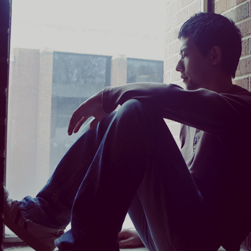 A monochromatic photo of Moro, a young Japanese man in a long-sleeved shirt and jeans sitting in a university window nook.