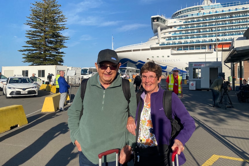 A man and a woman with luggage standing in front of a cruise ship