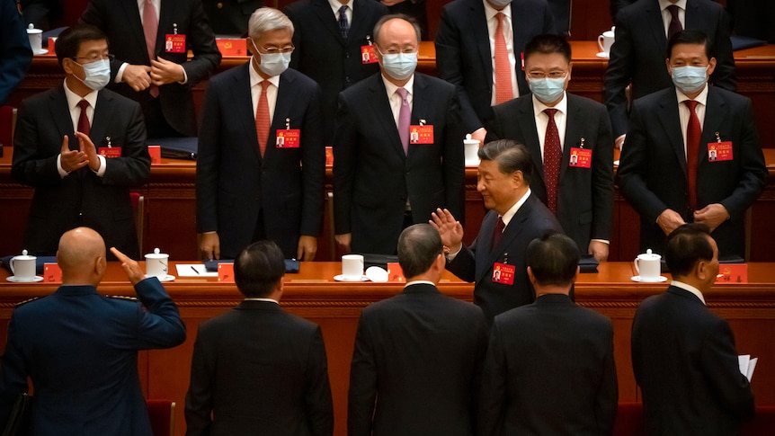 Rows of suited men stand and applaud as a leader walks down an aisle between them.