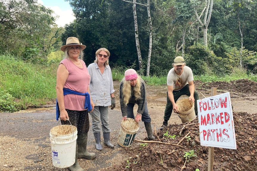 Three women and a man with buckets beside a pile of rubble woth a sign saying "follow marked paths".
