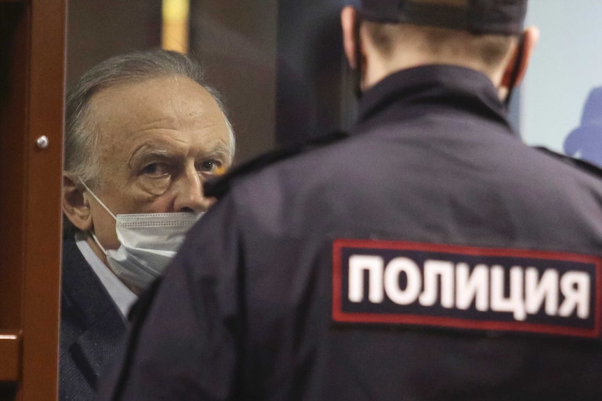 An elderly masked man looks pensive in court room watched by guard with Russian writing on his back.