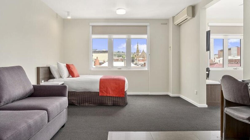 A spacious hotel room containing a couch, double bed, and other furniture. The view from the window is Hobart.