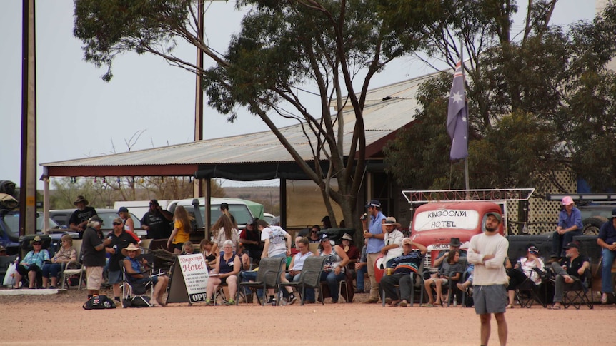 A large crowd sits on the red dirt under a metal verandah watching the cricket match.