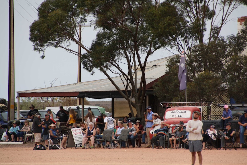 A large crowd sits on the red dirt under a metal verandah watching the cricket match.
