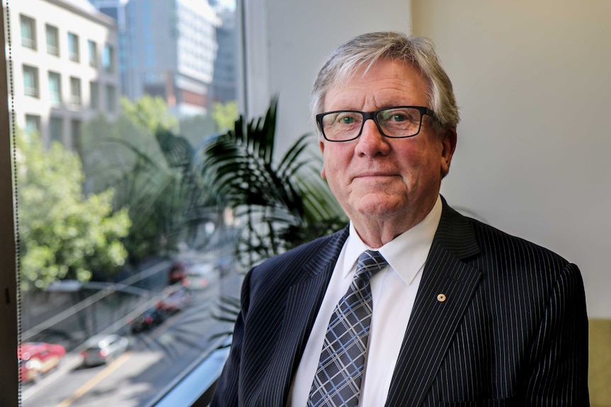 Phillip Cummins, wearing a suit and glasses, stands in an office room next to a window with a cityscape below.