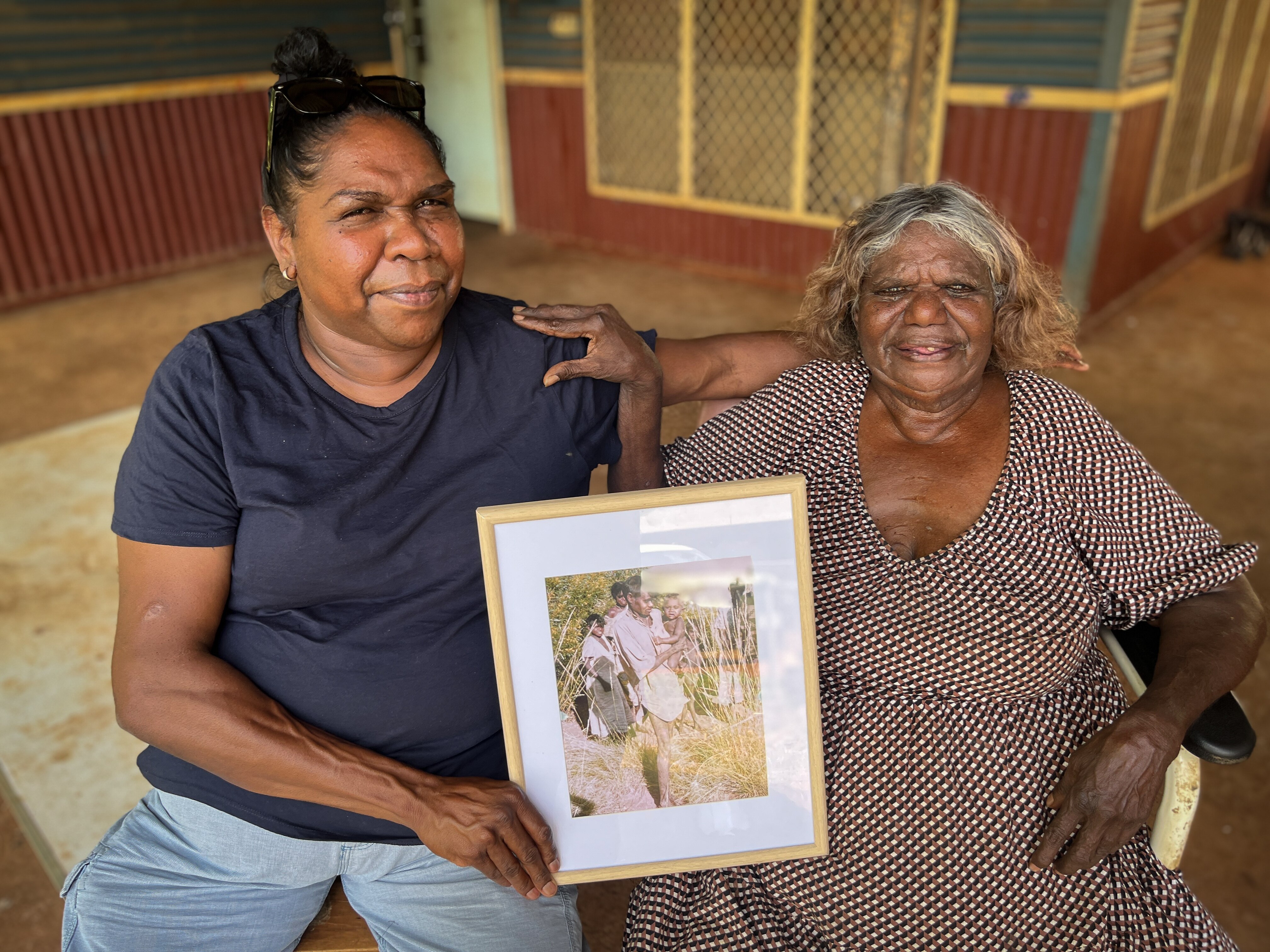Una mujer aborigen mayor sonríe y coloca una mano en el hombro de otra que sostiene una fotografía enmarcada.