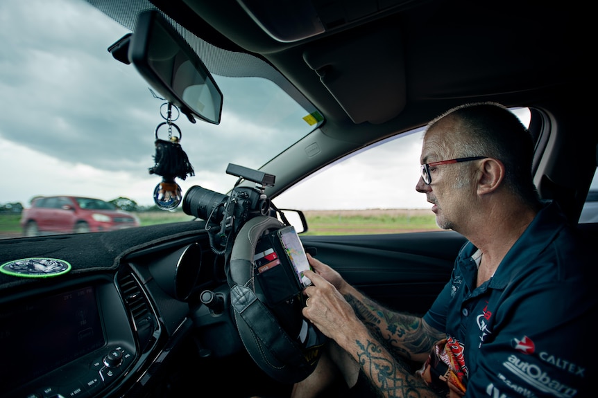 A man looks at his phone while sitting in the driver's seat of a car. A camera is propped up on the dashboard.