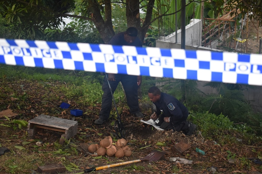 One man squats on ground while another holds metal detector behind police tape.