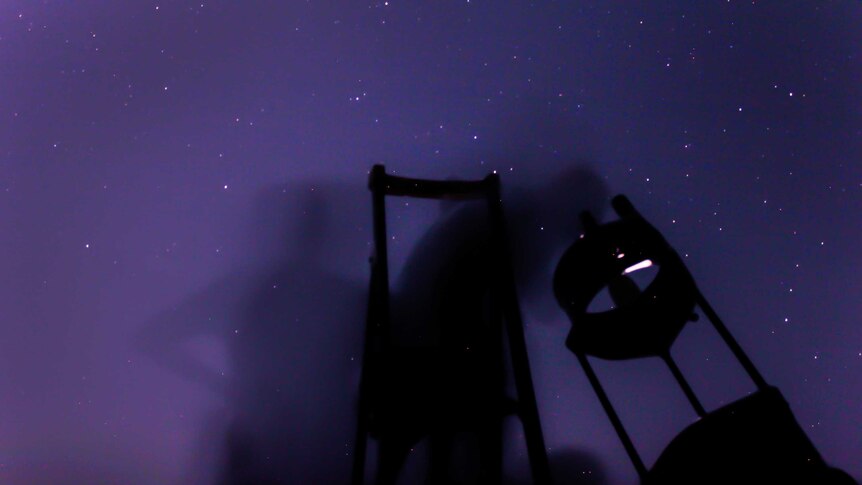 Blurry stargazers peer into one of the telescopes owned by the tour group with a purple starry background.