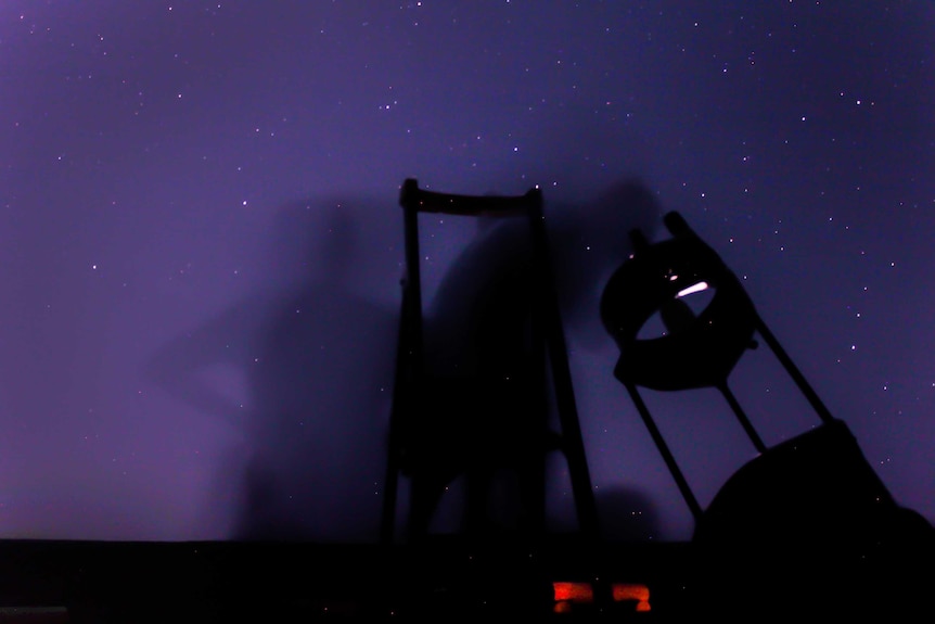 Blurry stargazers peer into one of the telescopes owned by the tour group with a purple starry background.