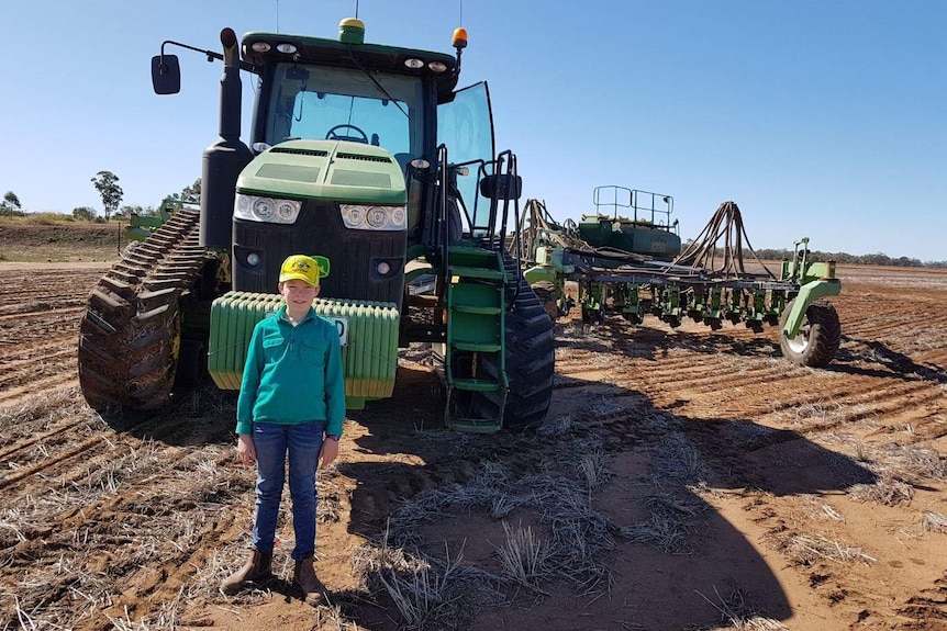 A boy stands in front of a large tractor on a farm.