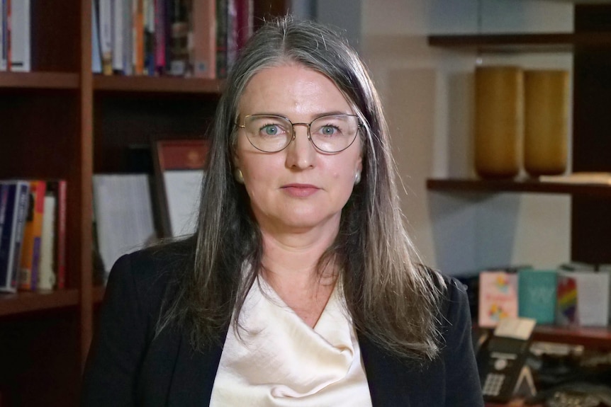 A woman wearing glasses,  black jacket and white top.