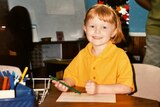 a young girl with red hair and yellow tshirt sits smilling a a desk holding a green texta