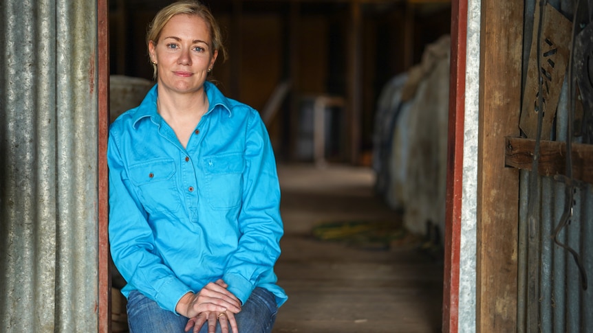 Woman in a blue long-sleeve shirt sits leaning against a shed entry way.