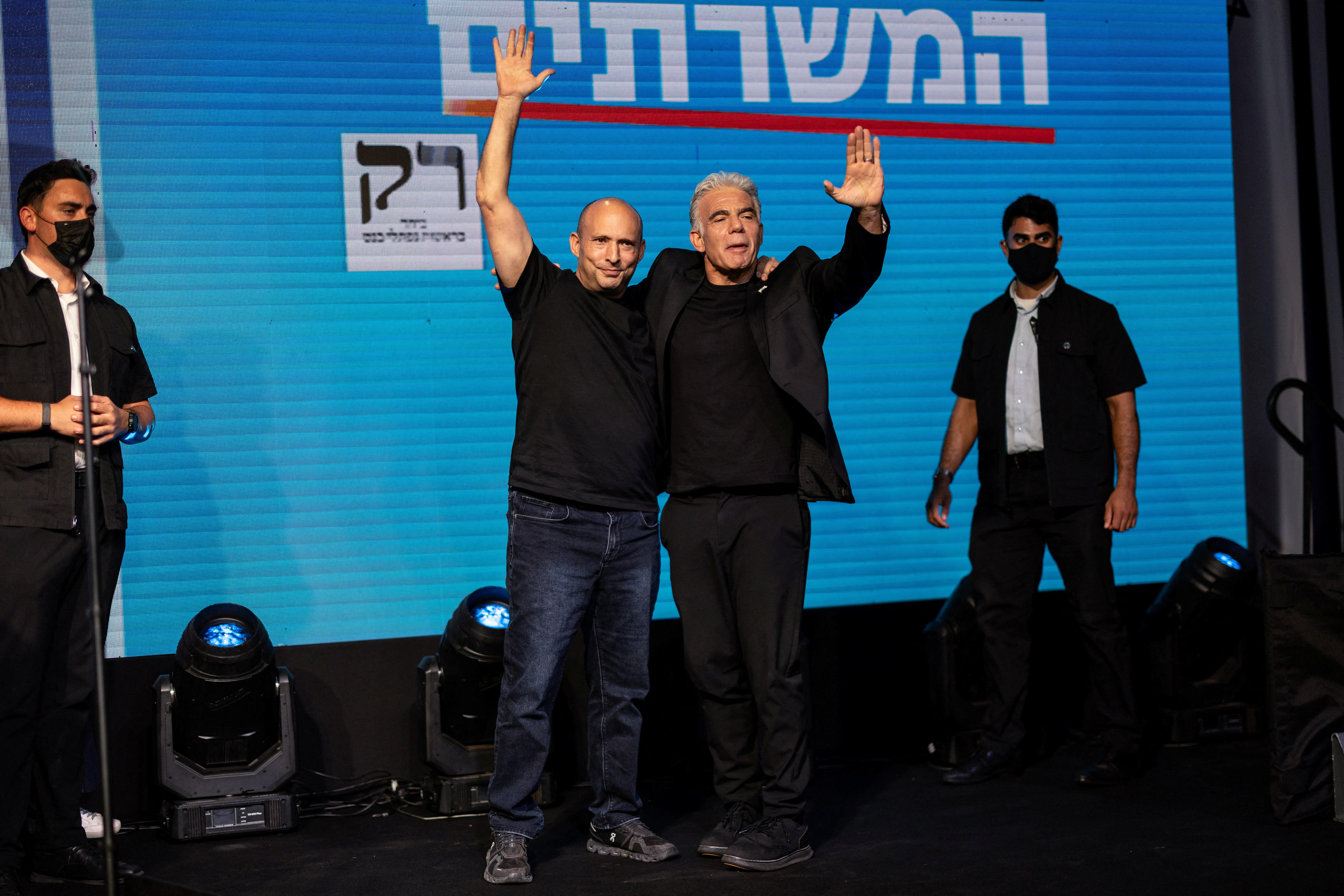 Naftali Bennett and Yair Lapid standing on stage with their arms wrapped around each other and hands up in the air