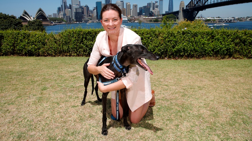 A smiling woman kneels down with her arms around a greyhound, with the Sydney Harbour Bridge and Sydney Opera House behind her.