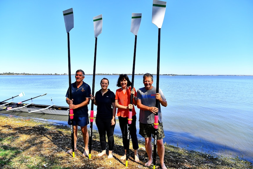 Four people stand holding oars in font of a body of water with a row boat to their right. 