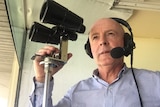 Greg Miles holds a fixed pair of binoculars and wears a headset with microphone.