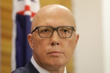 Peter Dutton wearing black glasses standing in front of a blurry australian flag looking straight toward the camera