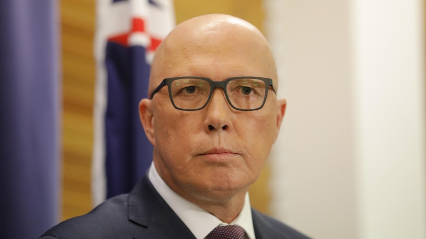 Peter Dutton wearing black glasses standing in front of a blurry australian flag looking straight toward the camera