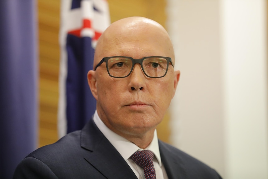 Peter Dutton wearing black glasses standing in front of a blurry australian flag looking straight toward the camera