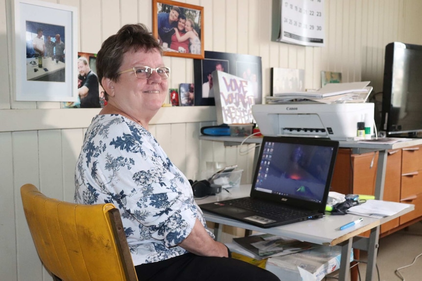 An older woman with short hair, glasses and a floral-print blouse sits in front of a laptop in a home office.