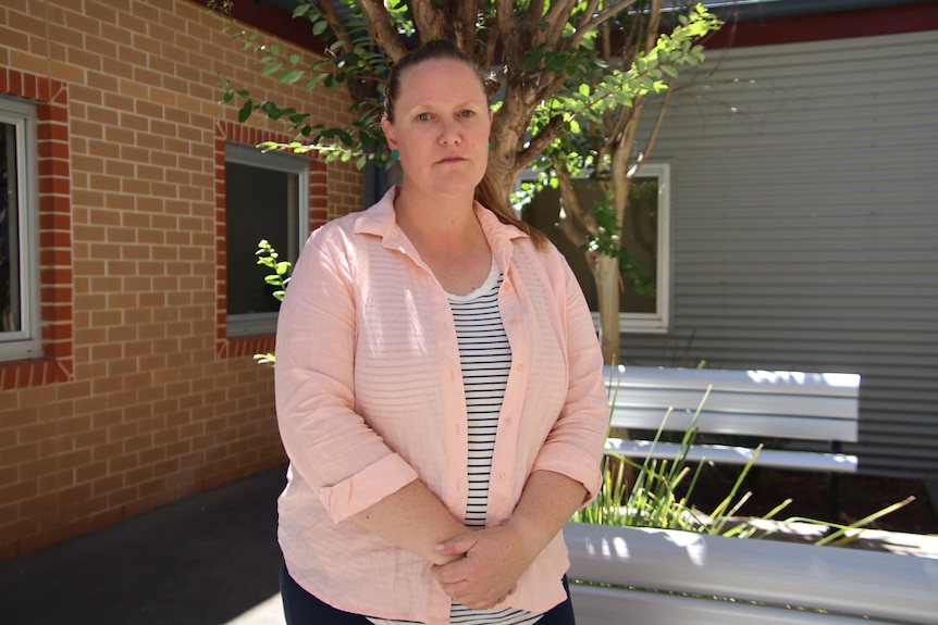 A woman wearing a striped shirt and pink blouse looking serious with hands clasped in front of a metal bench and tree