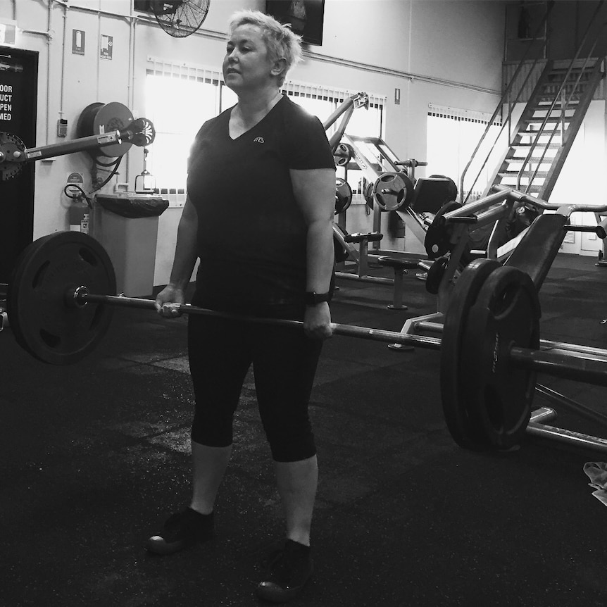 A middle-aged woman stands and holds weights beneath her waist.