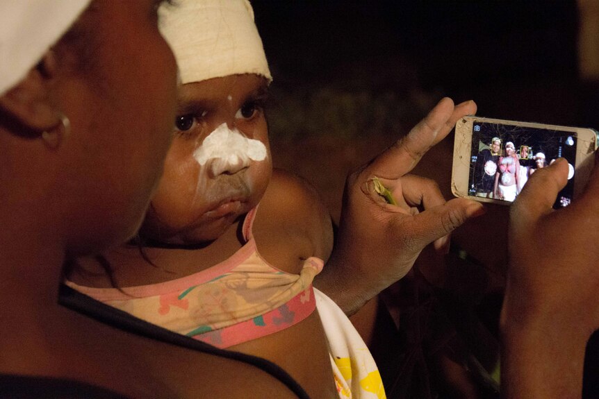 A baby looks on a woman takes a photo of people painted up for traditional Aboriginal dancing.