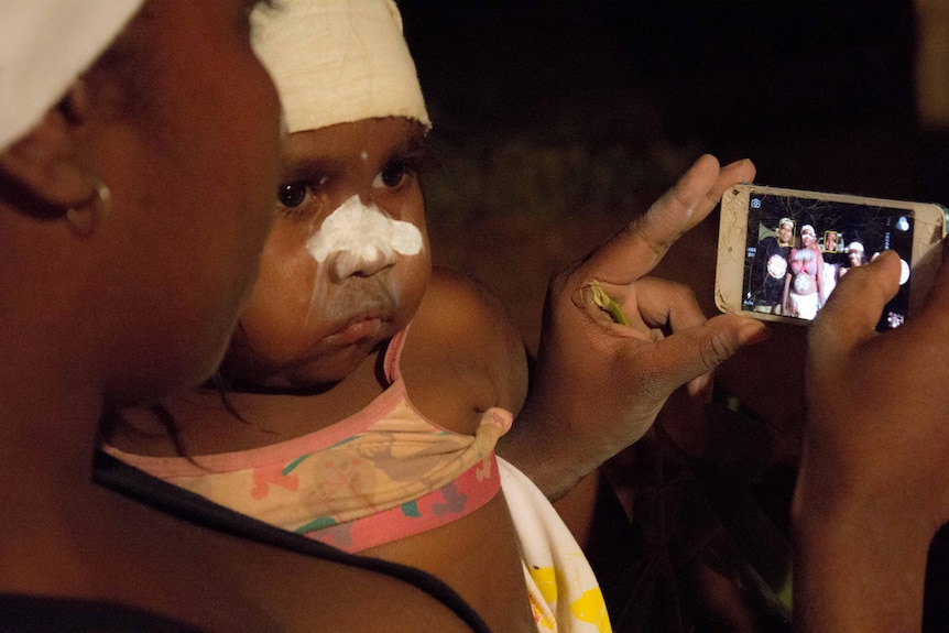 Ein Baby sieht eine Frau an, die ein Foto von Menschen macht, die für den traditionellen Tanz der Aborigines bemalt sind.