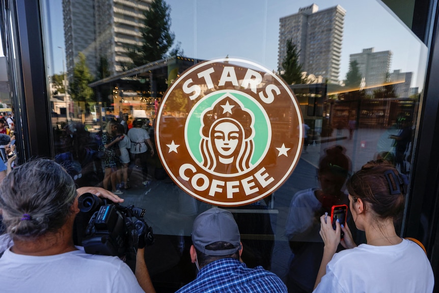 Cilvēki pulcējas ap apļa logotipu ar uzrakstu Stars Coffee pie kafejnīcas skatloga.