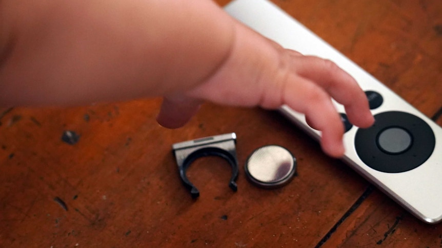 A six-month-old baby reaches for an Apple TV remote, with a button battery beside