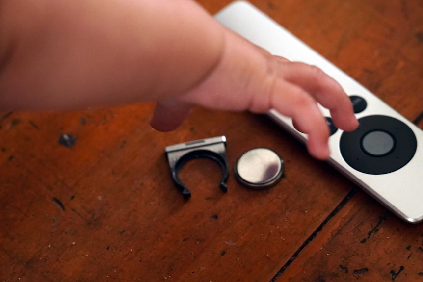 A six-month-old baby reaches for an Apple TV remote, with a button battery beside