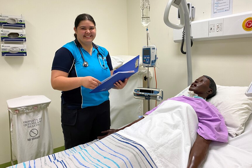 Nursing student in blue shirt attends to mock patient in hospital bed