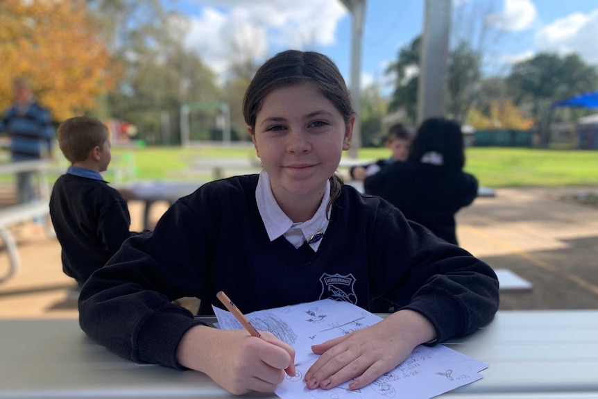 School girl sitting at outdoor table drawing on piece of paper.