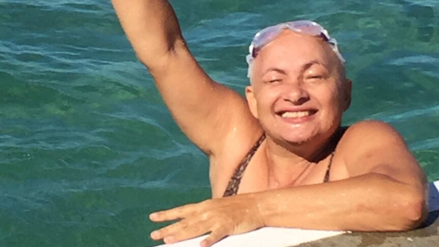 A woman with short grey hair in an ocean pool raises her arm out of the water to give the peace sign