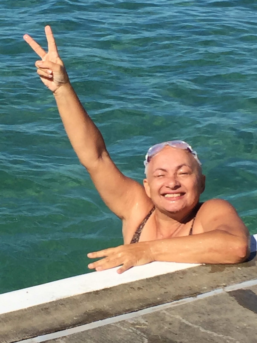 A woman with short grey hair in an ocean pool raises her arm out of the water to give the peace sign