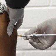 The shoulder of a volunteer is seen as they receive an injection by a person wearing gloves.