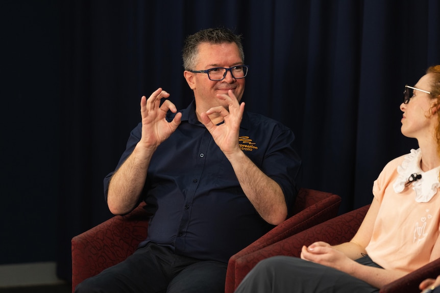 A man in a navy polo shirt displays sign language sitting in a chair while a woman watches on.