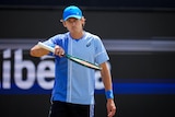 A male tennis player on court, holding a racket, wearing light blue shirt and cap.