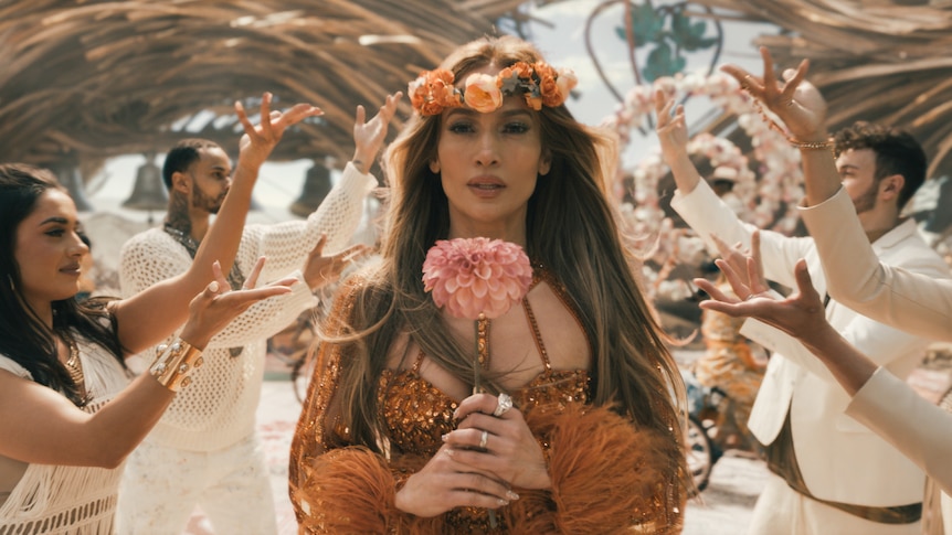 Jennifer Lopez wears a flower garland and carries a bouquet as she walks down an aisle surrounded by dances forming an arch.