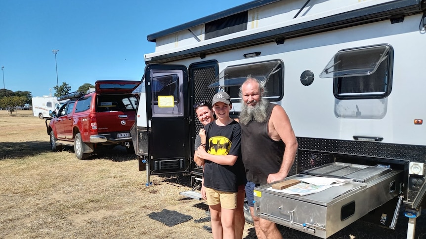 Three smiling people stand in front of a caravan in the outback.