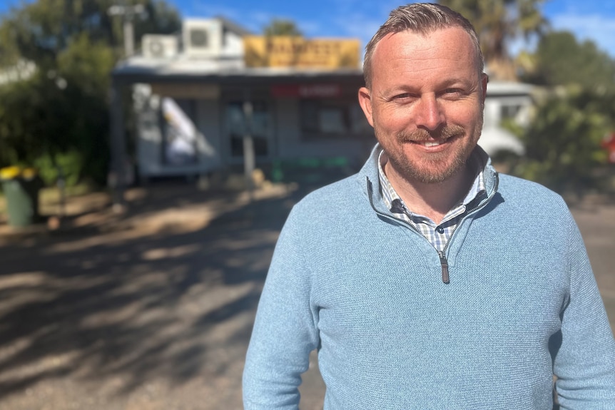 A smiling man with short, fair stands outside a small building, smiling in the sun.