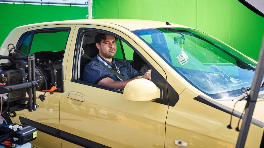 Bjorn Stewart dressed as a policeman looks out the window of a yellow hatchback during the filming of a TV series KGB