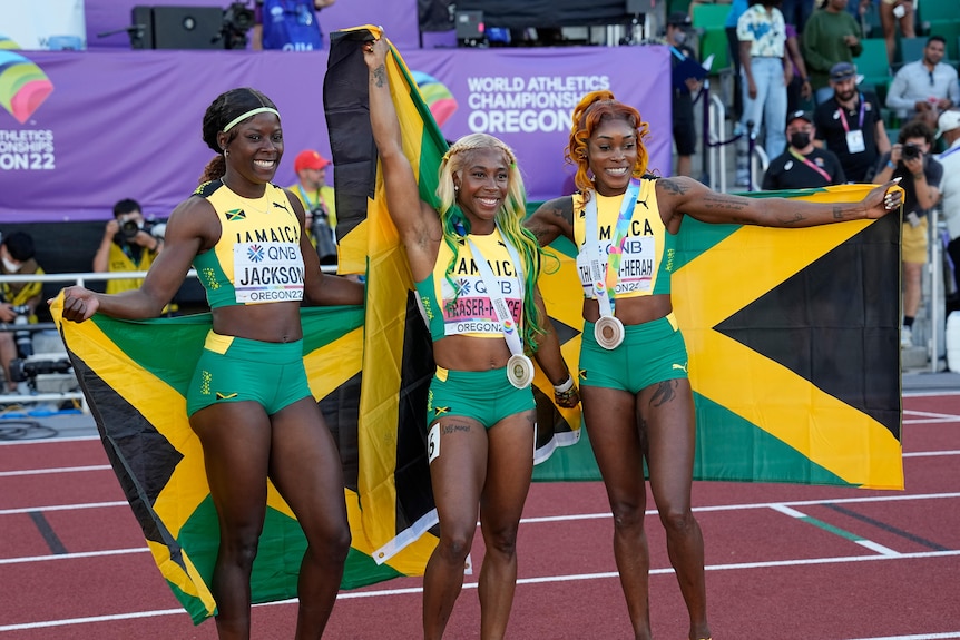 Three women sprinters pose with Jamaican flags and medals, smiling.