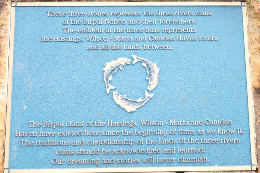 A blue plaque on a stone featuring three bass fish forming a circle that represent 'the three river clans of the Birpai Nation.'