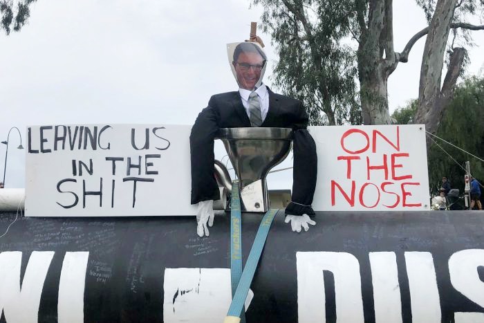 An effigy in a black suit with a picture of mans face sits in toilet next to protest signs