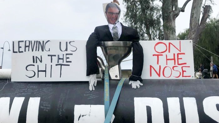 An effigy in a black suit with a picture of mans face sits in toilet next to protest signs.