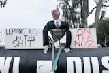 An effigy in a black suit with a picture of mans face sits in toilet next to protest signs.