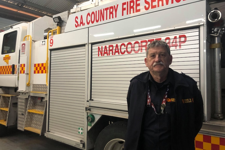 A man with a moustache stands in front of a firetruck